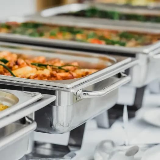 A row of professional stainless steel catering chafing dishes filled with gourmet food on a white tablecloth, representing luxury catering services.