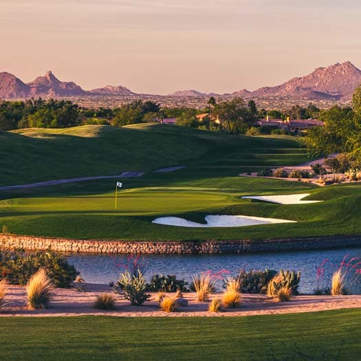 A scenic desert golf course at golden hour, with a green, water, and mountains beyond.