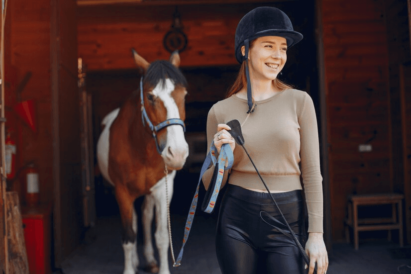 Smiling woman in riding gear leads a horse out of a stable.