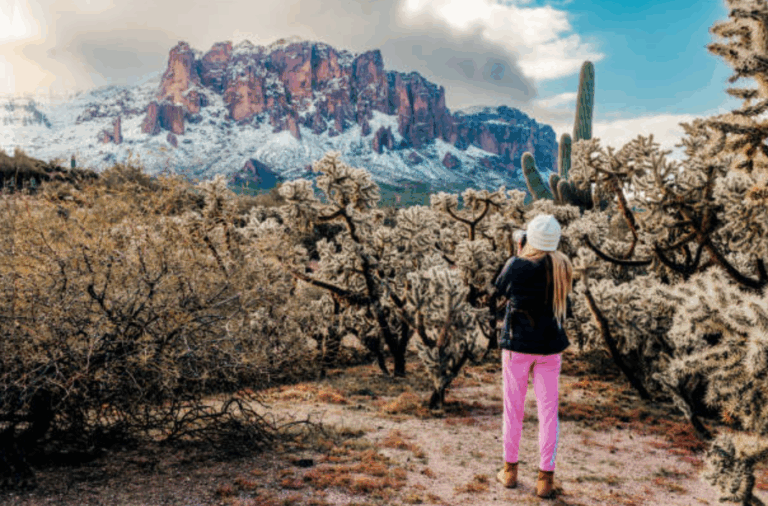 Snow-covered Superstition Mountains with cacti in the foreground and a woman taking photos.