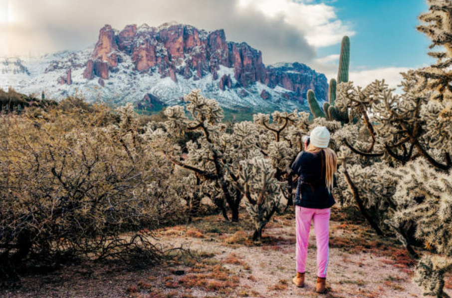 Snow-covered Superstition Mountains with cacti in the foreground and a woman taking photos.